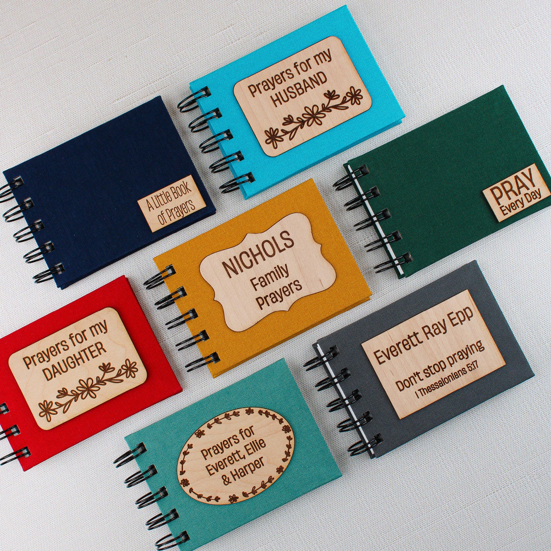 Small spiral prayer books in colorful bookcloth with wooden plaques featuring family prayers, personal names and prayer titles.