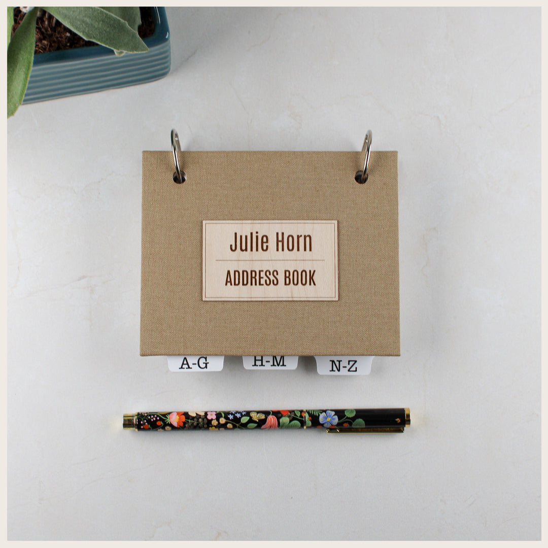 Khaki book cloth address book with a personalized rectangular title plate and alphabetical tabs, shown above a floral pen.