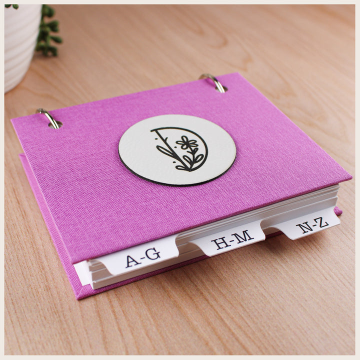 Pink book cloth password keeper book with alphabetical tabs and a floral D initial on a round white nameplate.