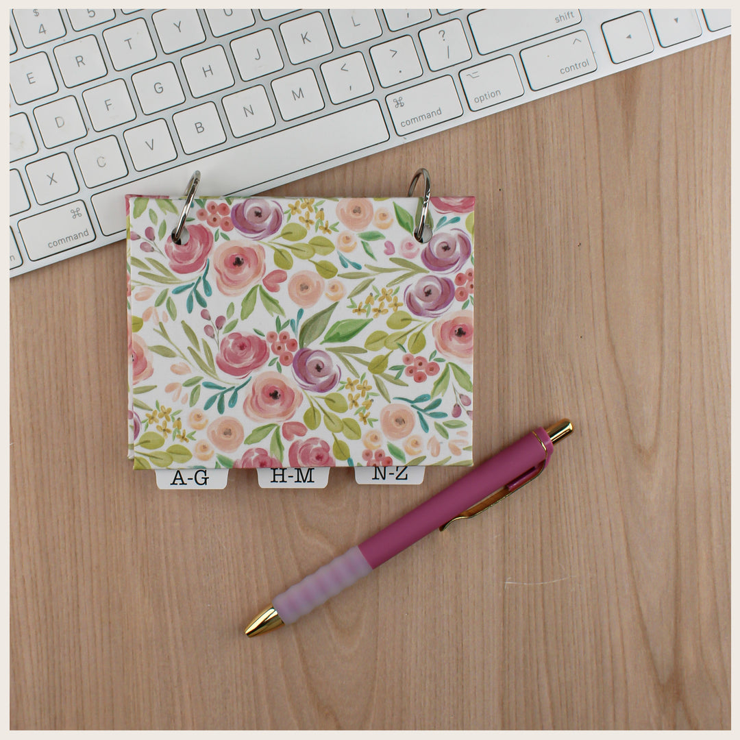 Pink and green floral fabric password book with a blank cover and alphabetical tabs, beside a keyboard and pink pen.