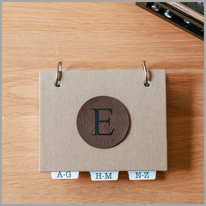 Desk accessory password book in khaki bookcloth with dark brown leatherette initial E in a 2" circle affixed to front cover.