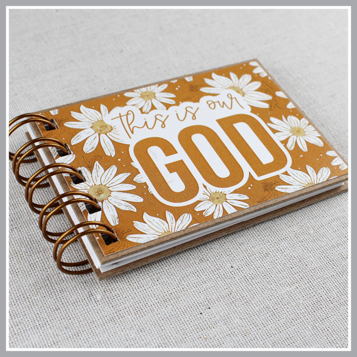 Angled close up of small spiral bound attributes of God book in Daisy style with yellow background and ivory flowers. Front cover says "this is our GOD".