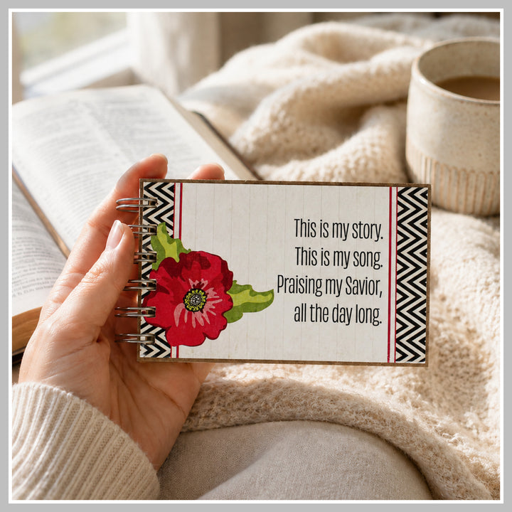 Red flower Christian prayer journal held in hand beside a Bible and coffee.