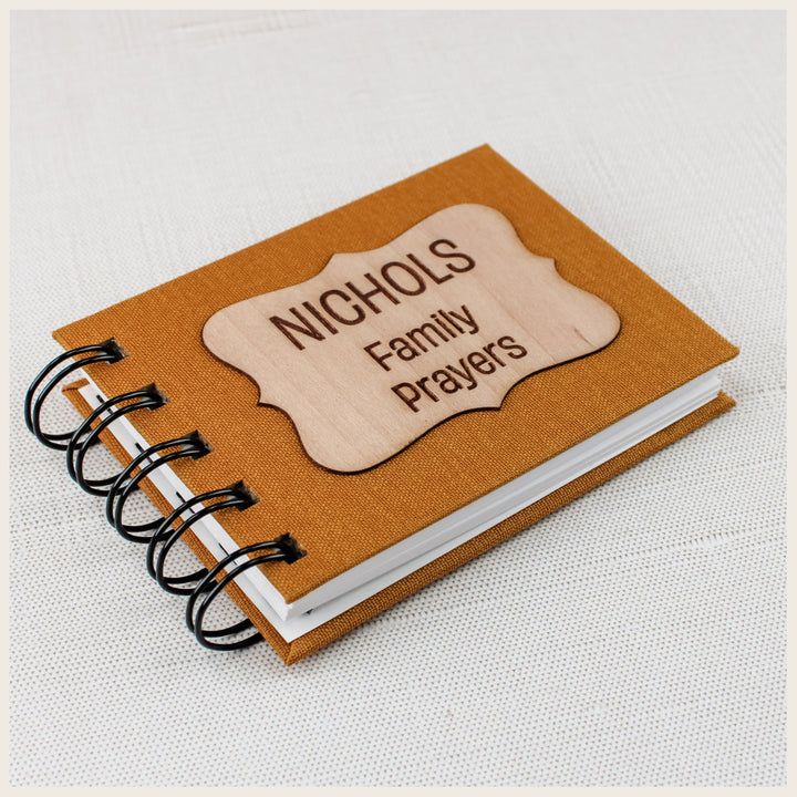 Angled close-up of a mustard family prayer book showing its NICHOLS Family Prayers wood plaque, cloth hardcover and spiral binding.