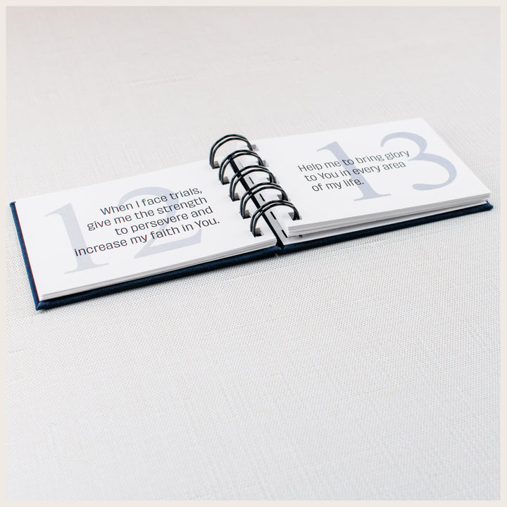 Angled view of an open spiral prayer book showing prayers 12 and 13 about persevering through trials and bringing glory to God.