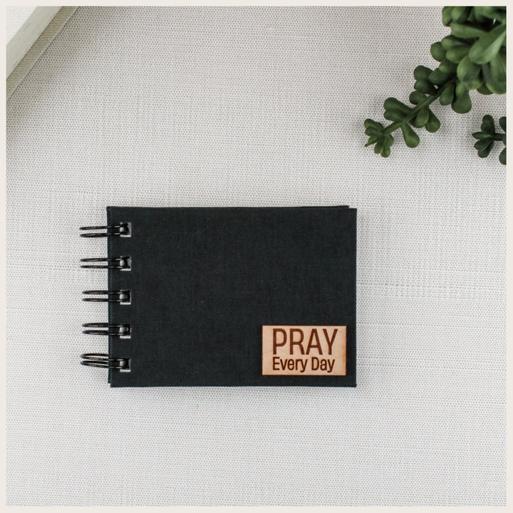 Black book cloth prayer book with spiral binding and a small wood plaque engraved PRAY Every Day.