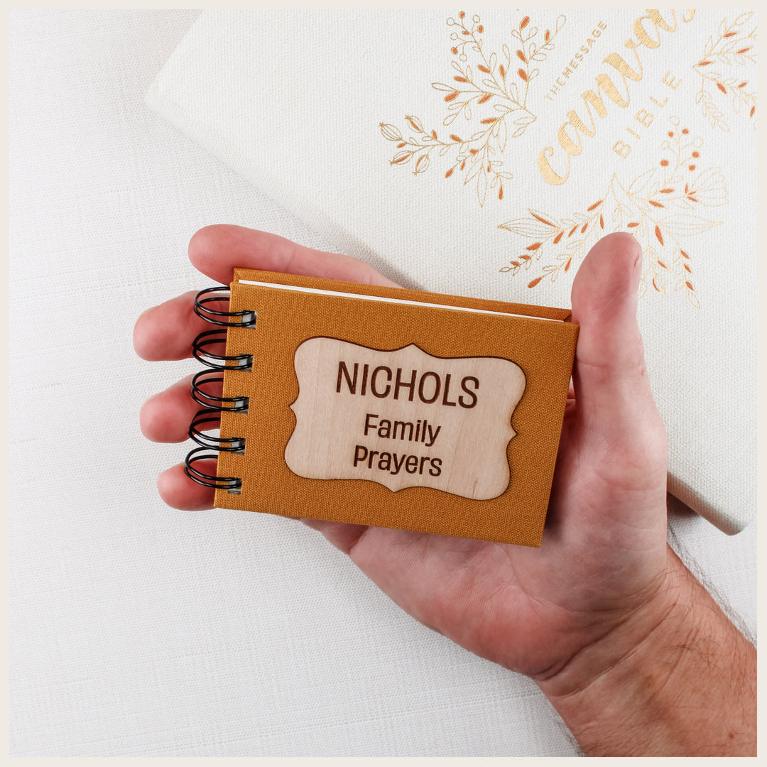 Small mustard book cloth prayer book resting in an adult’s palm, with a curly wood plaque engraved NICHOLS Family Prayers.