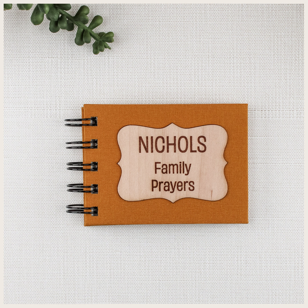 Mustard book cloth family prayer book with black spiral binding and a curly wood plaque engraved NICHOLS Family Prayers.
