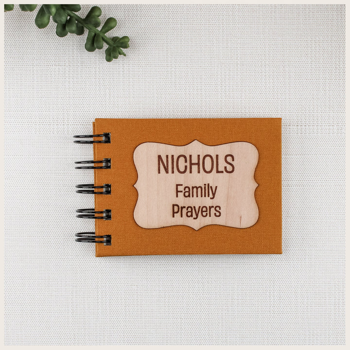 Mustard book cloth family prayer book with black spiral binding and a curly wood plaque engraved NICHOLS Family Prayers.