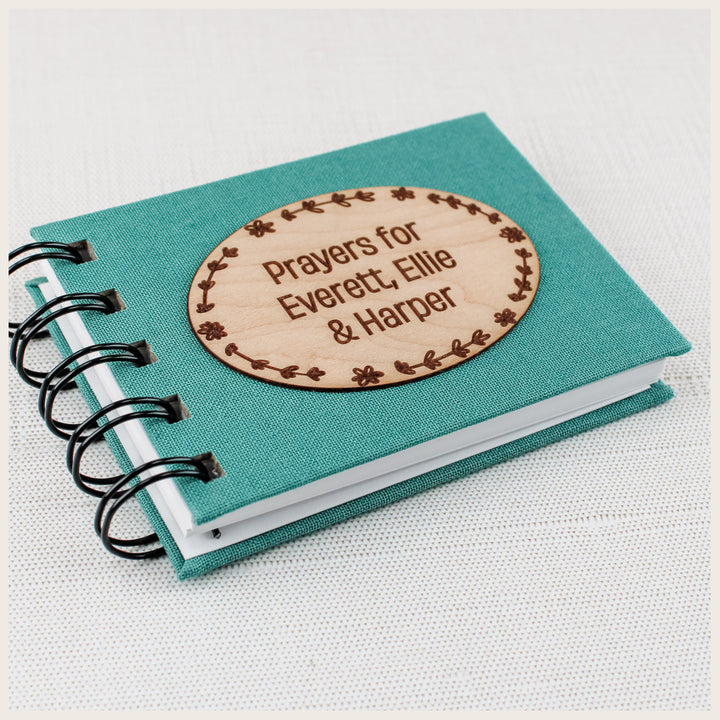 Close-up of a teal cloth prayer book with black spiral binding and a personalized floral oval wood plaque naming three children.