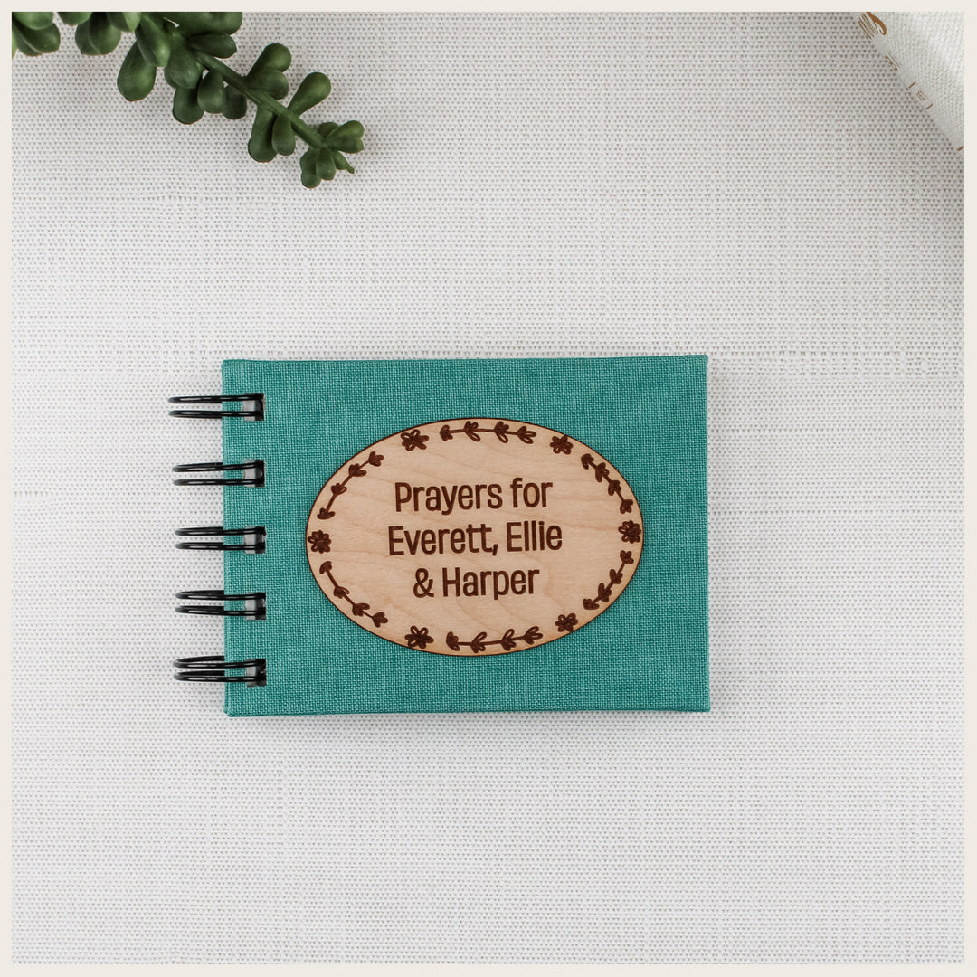 Teal cloth prayer book with a floral oval wood plaque engraved Prayers for Everett, Ellie &amp; Harper.