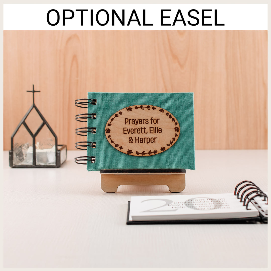 Personalized teal prayer book displayed on the optional wooden easel beside a church-shaped lantern.