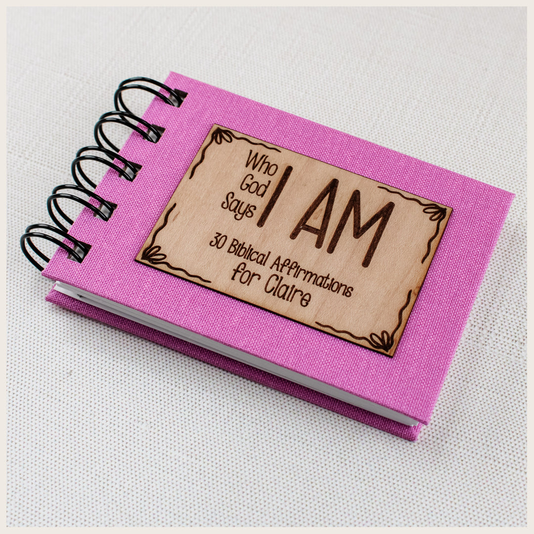 Close-up of pink book cloth, black spiral binding, and a bows wood plaque personalized for Claire on a Christian affirmation book.