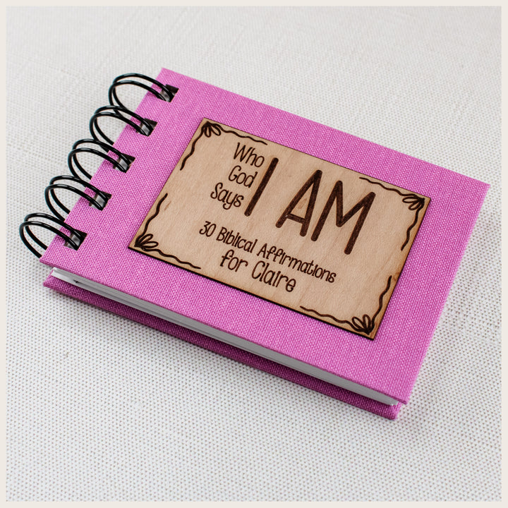 Close-up of pink book cloth, black spiral binding, and a bows wood plaque personalized for Claire on a Christian affirmation book.