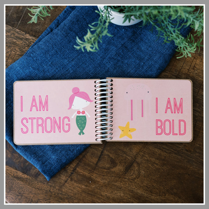 Open Pink girly affirmation book showing “I am strong” and “I am bold” pages with pink background and girly graphics. Mermaid, jelly fish and star fish on this page spread.