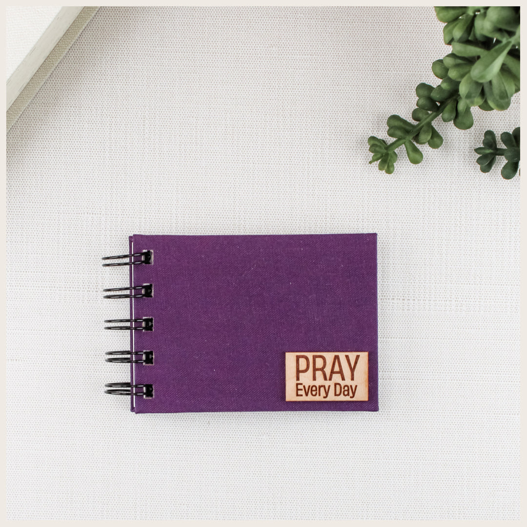 Plum cloth prayer book with black spiral binding and the included small wood plaque reading PRAY Every Day.