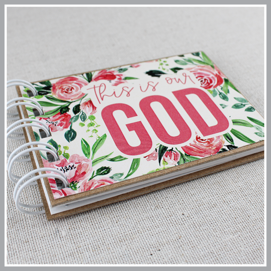 Close up photo at an angle to show details of attributes of God spiral bound book with pink flowers and green leaves. Front cover says "this is our GOD".
