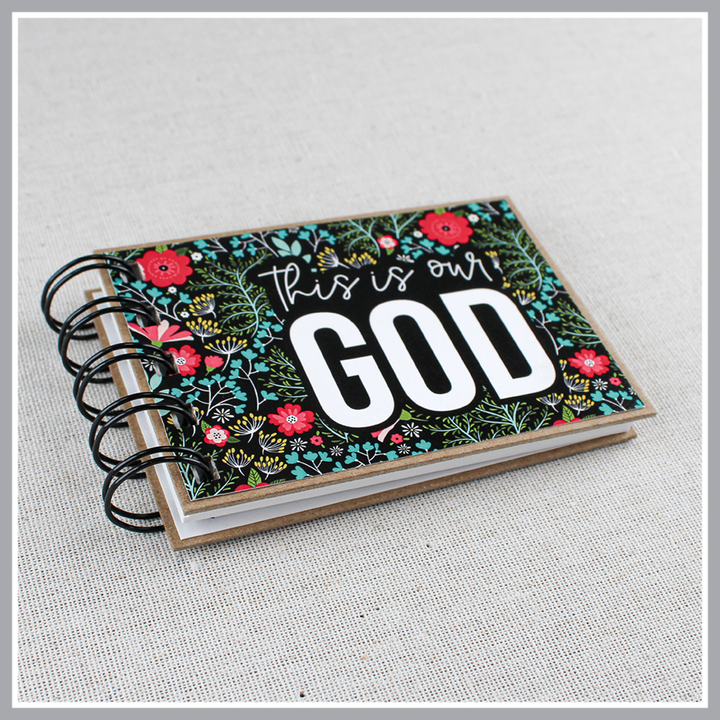 Angled, close up of God's attribute book with a black background and bright florals. The cover of this small, spiral-bound book says "this is our GOD".