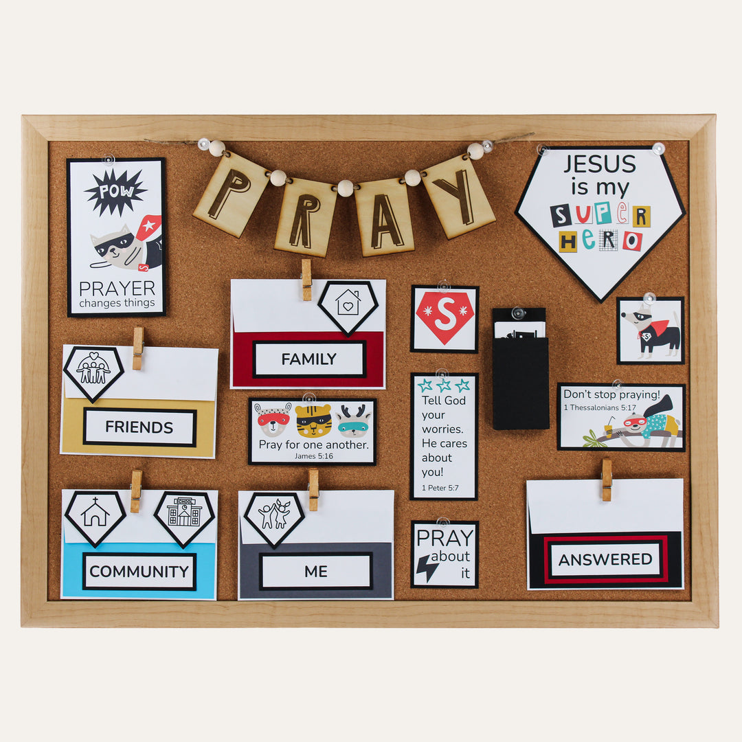 Superhero prayer board kit arranged on a corkboard with a wooden PRAY banner and Family, Friends, Community, Me, and Answered envelopes.