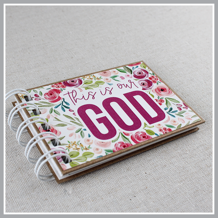 Angled close up photo of God's attributes book spiral bound in Pastel Floral style with pink and purple floral background. Front cover says "this is our GOD". 