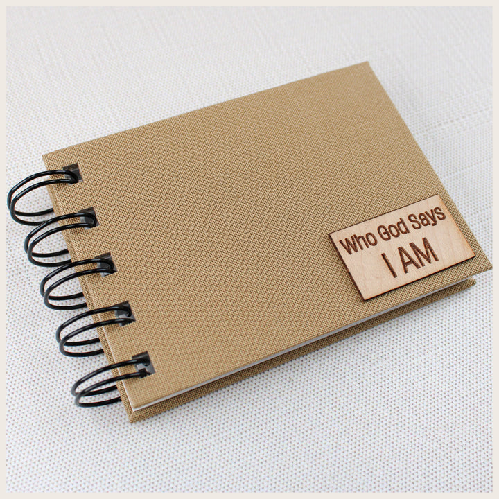 Close-up of a khaki spiral affirmation book with a small Who God Says I AM wood plaque in the lower right corner.