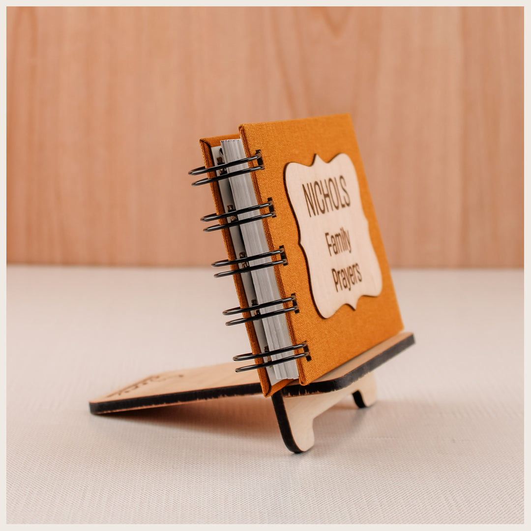 Side view of a wood easel supporting a gold spiral family prayer book at a backward angle. Book sold separately.
