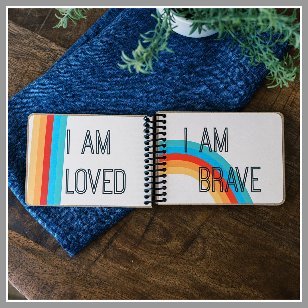 Open Rainbow affirmation book showing “I am loved” and “I am brave” pages with ivory background and rainbow graphics with a retro feel.